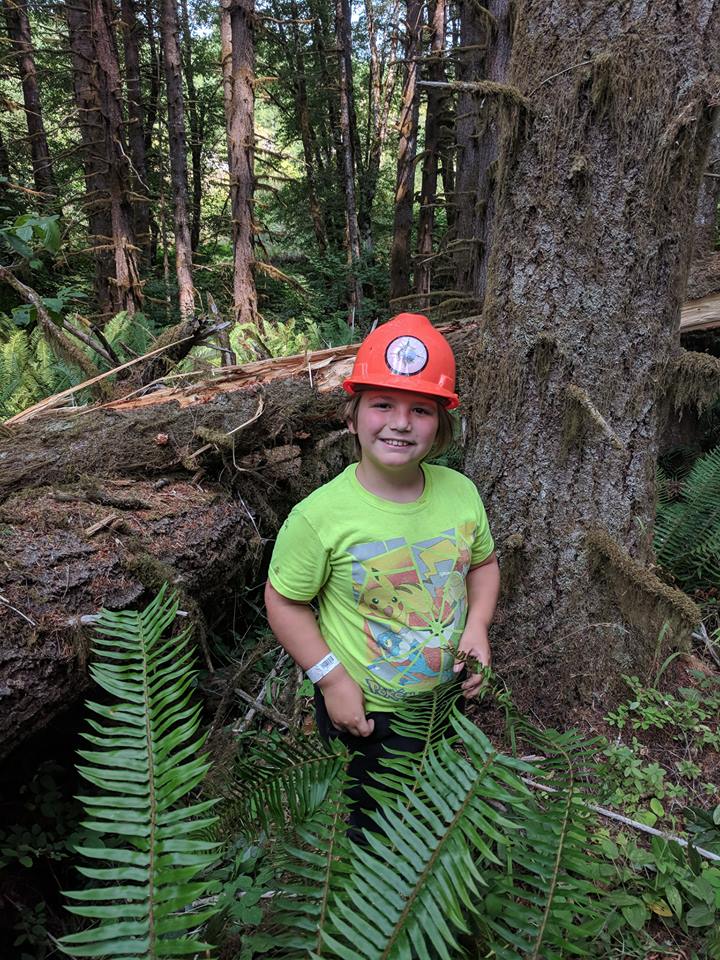 A boy standing in front of a tree, wearing an orange construction hat and a green shirt.
